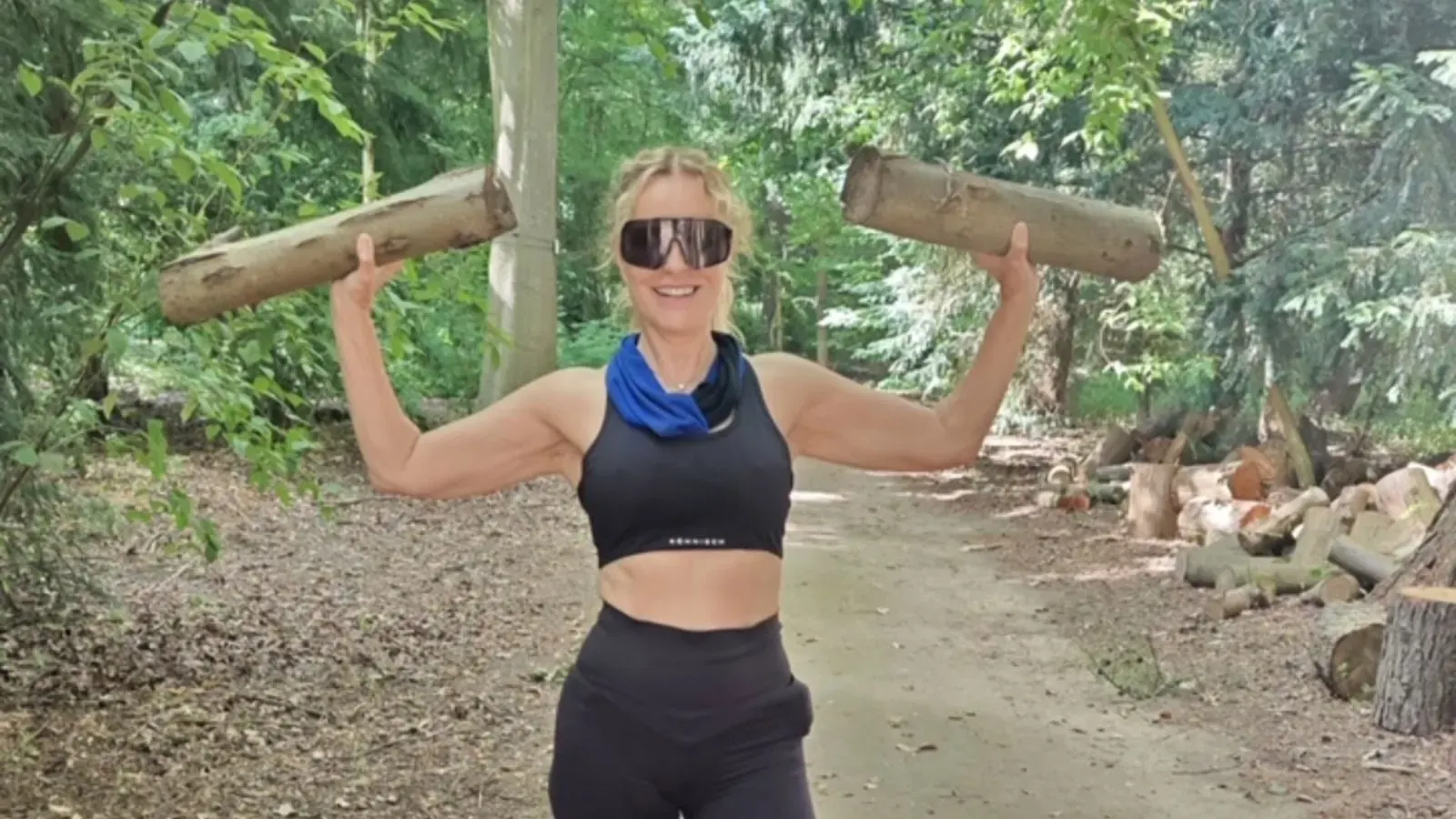 Katja Burkard hebt im Wald mit einem Sport-BH bekleidet Holzstämme über ihren Kopf.